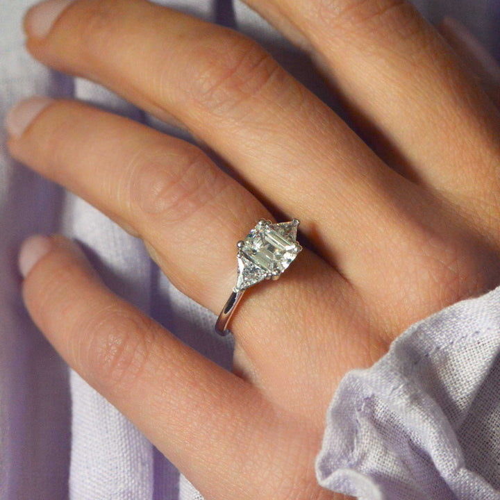 Close-up of a hand wearing an emerald cut diamond trilogy engagement ring with a soft background