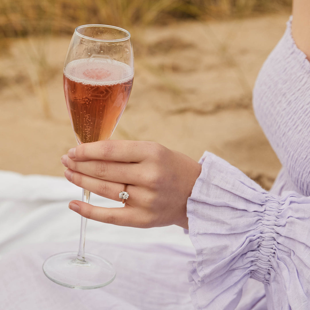 Hand holding a glass of pink champagne , wearing an oval cut engagement ring with a blurred natural background