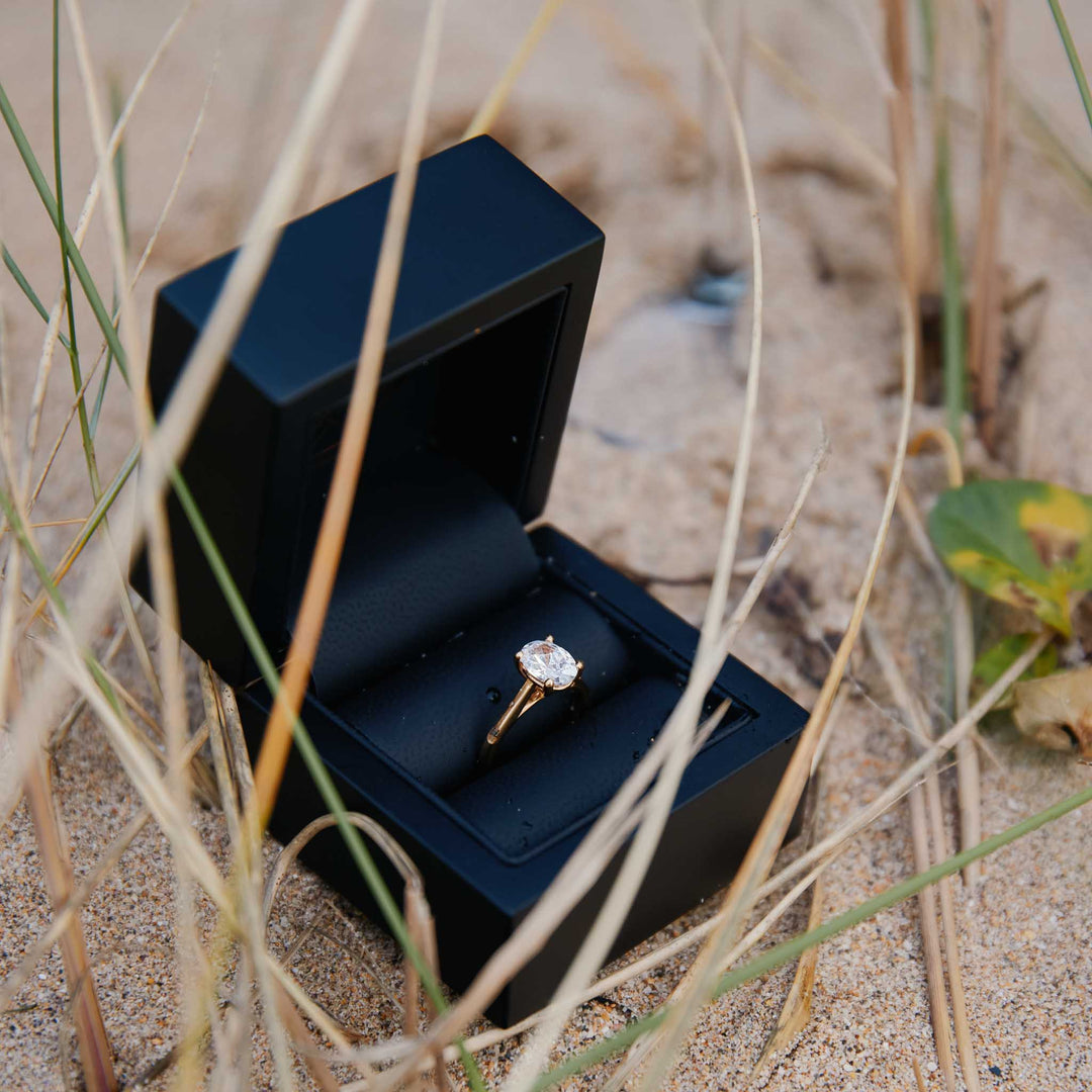 Oval diamond ring in a black box on sand after a beach proposal in Cornwall