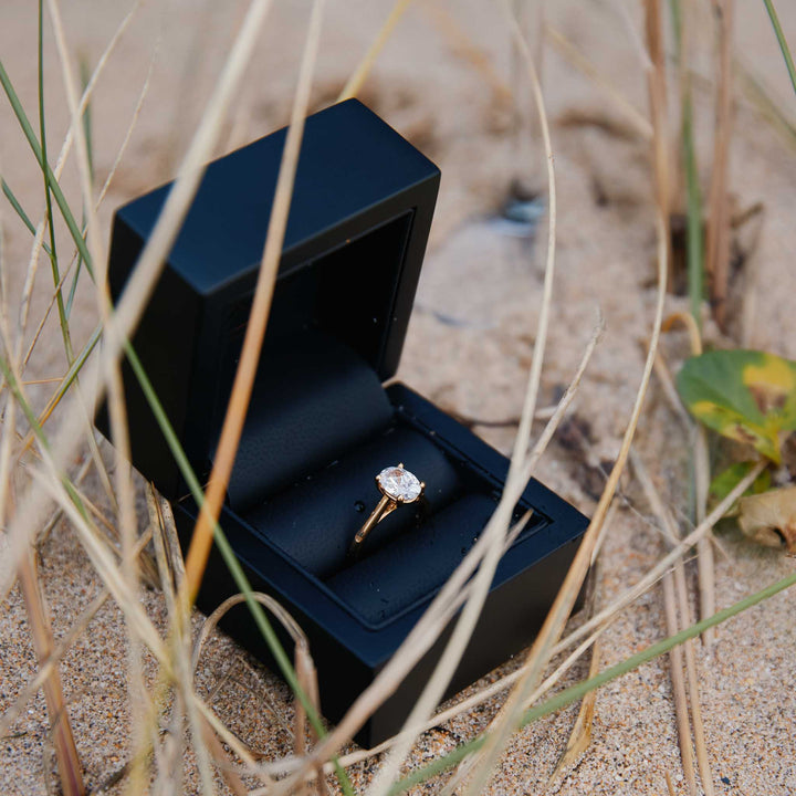 Oval diamond ring in a black box on sand after a beach proposal in Cornwall