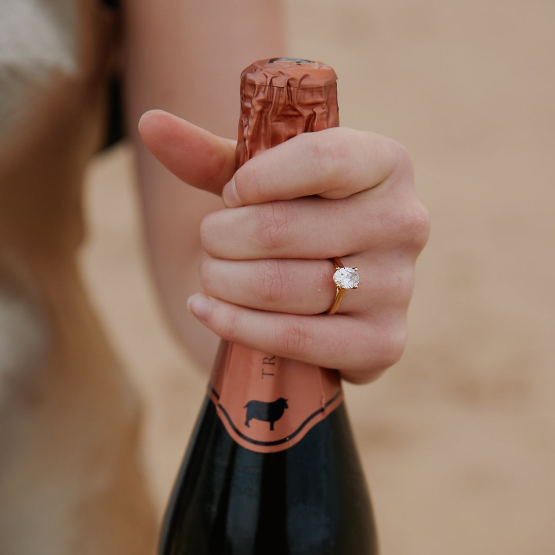 Hand wearing a oval diamond ring, holding a bottle of champagne against a blurred background