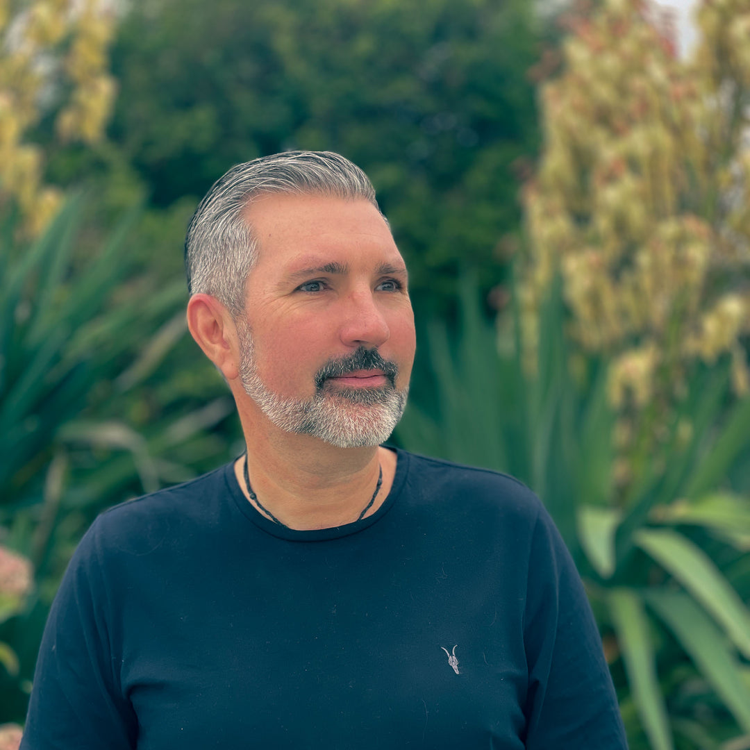 Man with gray hair and beard wearing a dark blue shirt standing in front of green plants.