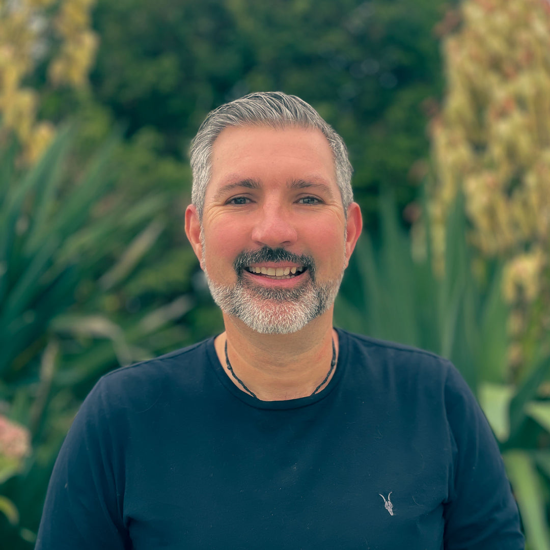 Man with gray hair and beard wearing a dark blue shirt in front of green foliage