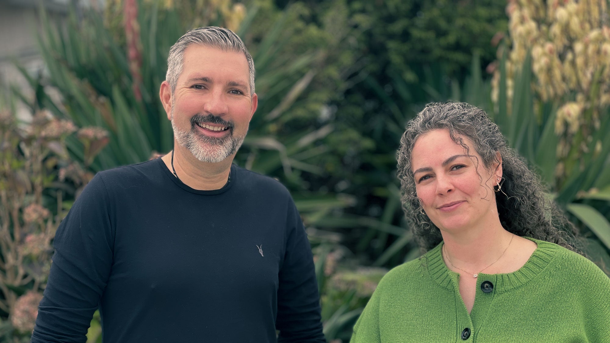 Two people standing outdoors with greenery in the background