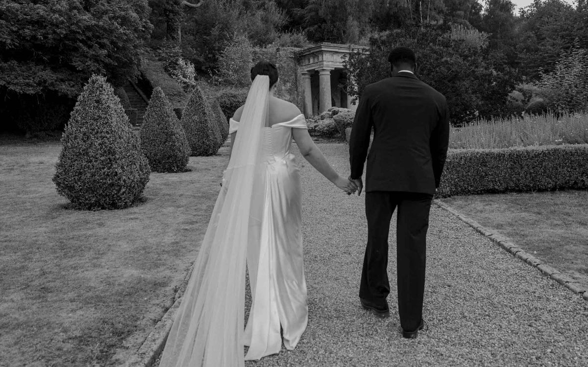 A black and white photograph of a bride in a Vivienne Westwood wedding dress with a long veil and a Groom in a dark suit walking hand-in-hand down a garden path toward a stone pavilion.