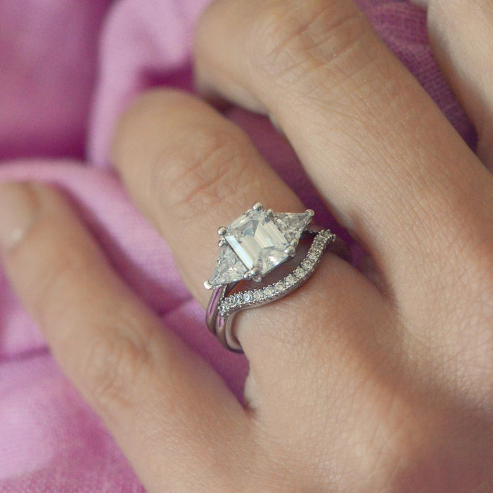 Platinum trilogy ring with a large emerald cut diamond, worn on the finger with a curved pavé wedding band on a hand against a pink fabric background