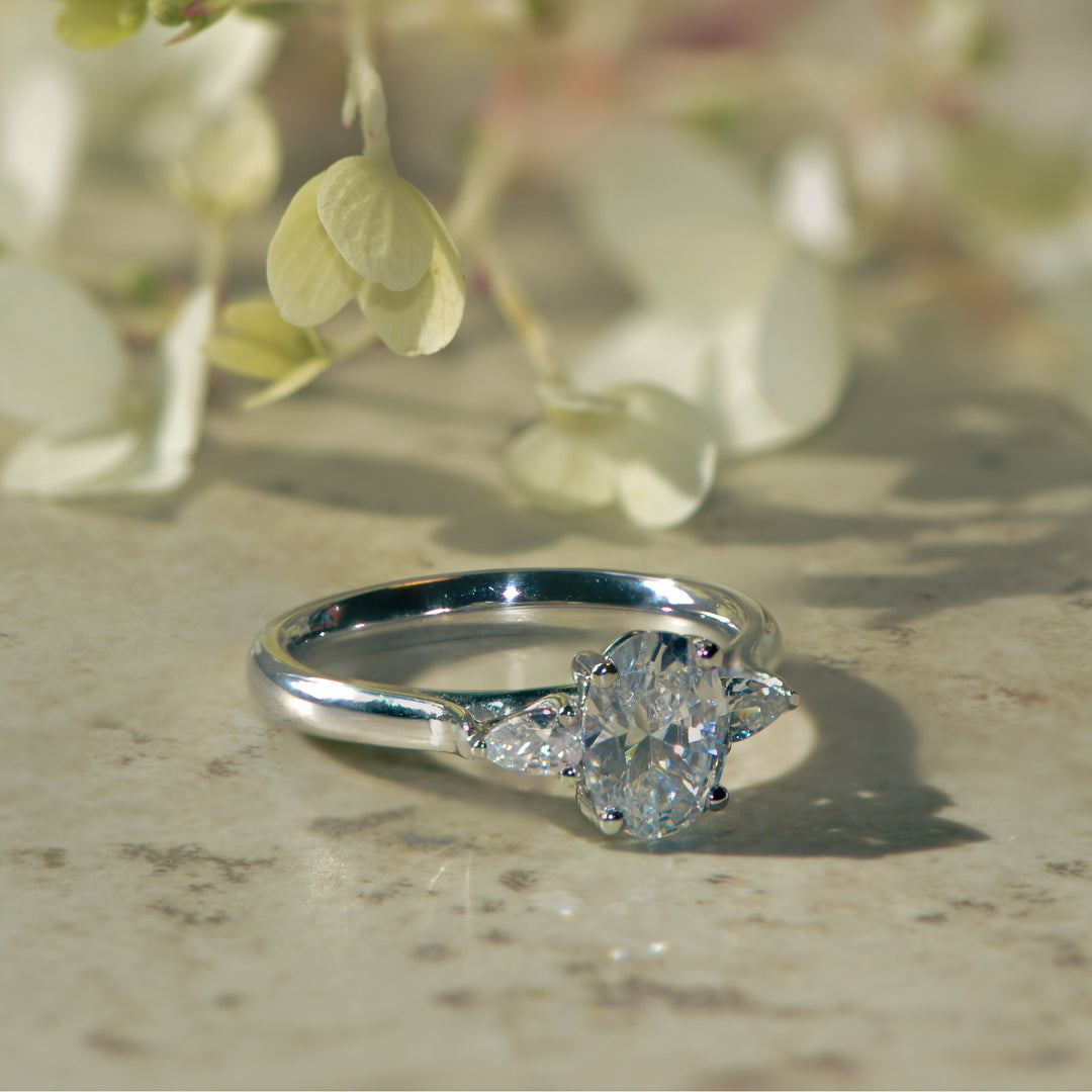 Oval cut trilogy diamond ring on a textured surface with greenery