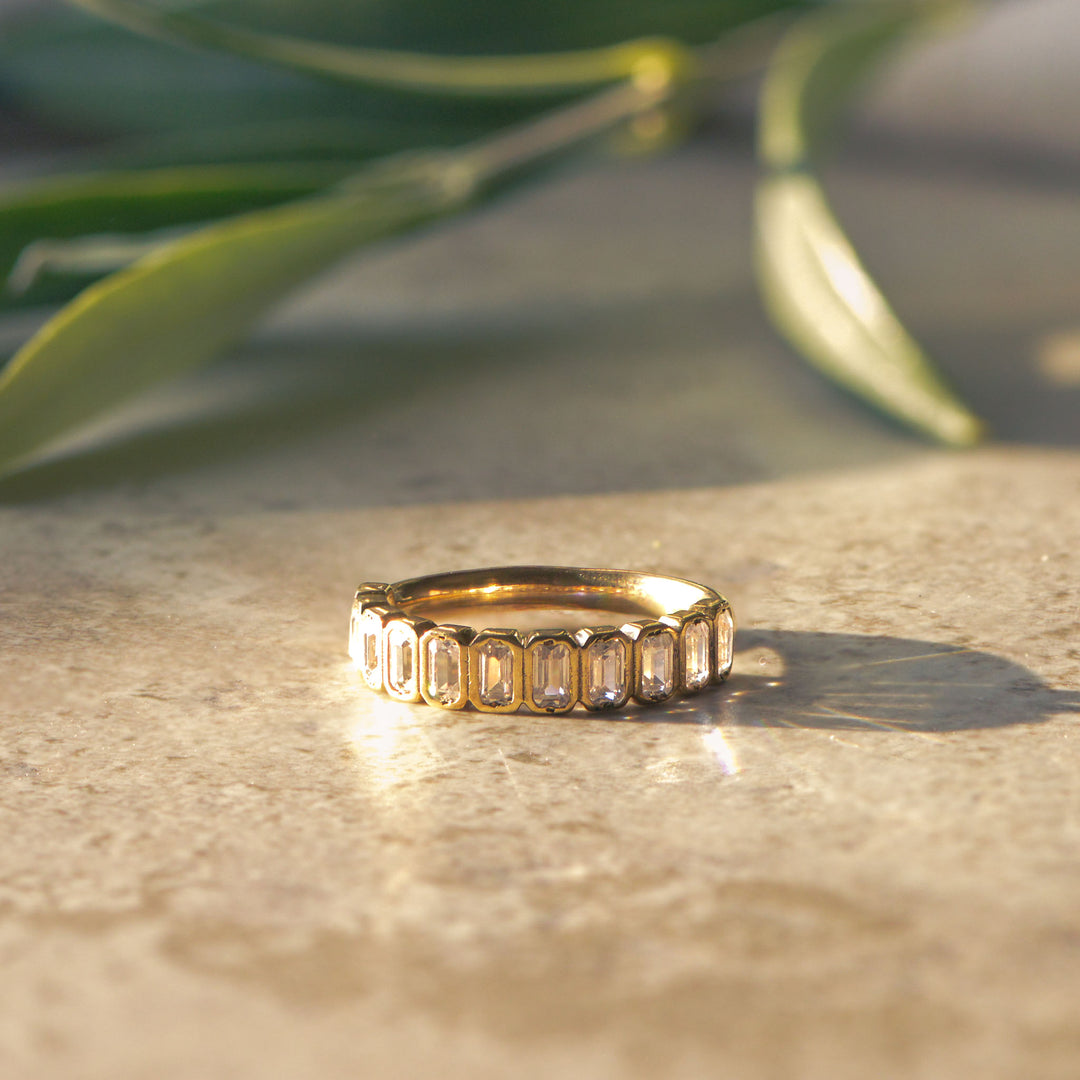 Gold wedding band bezel set with emerald cut diamonds on a stone surface with greenery in the background