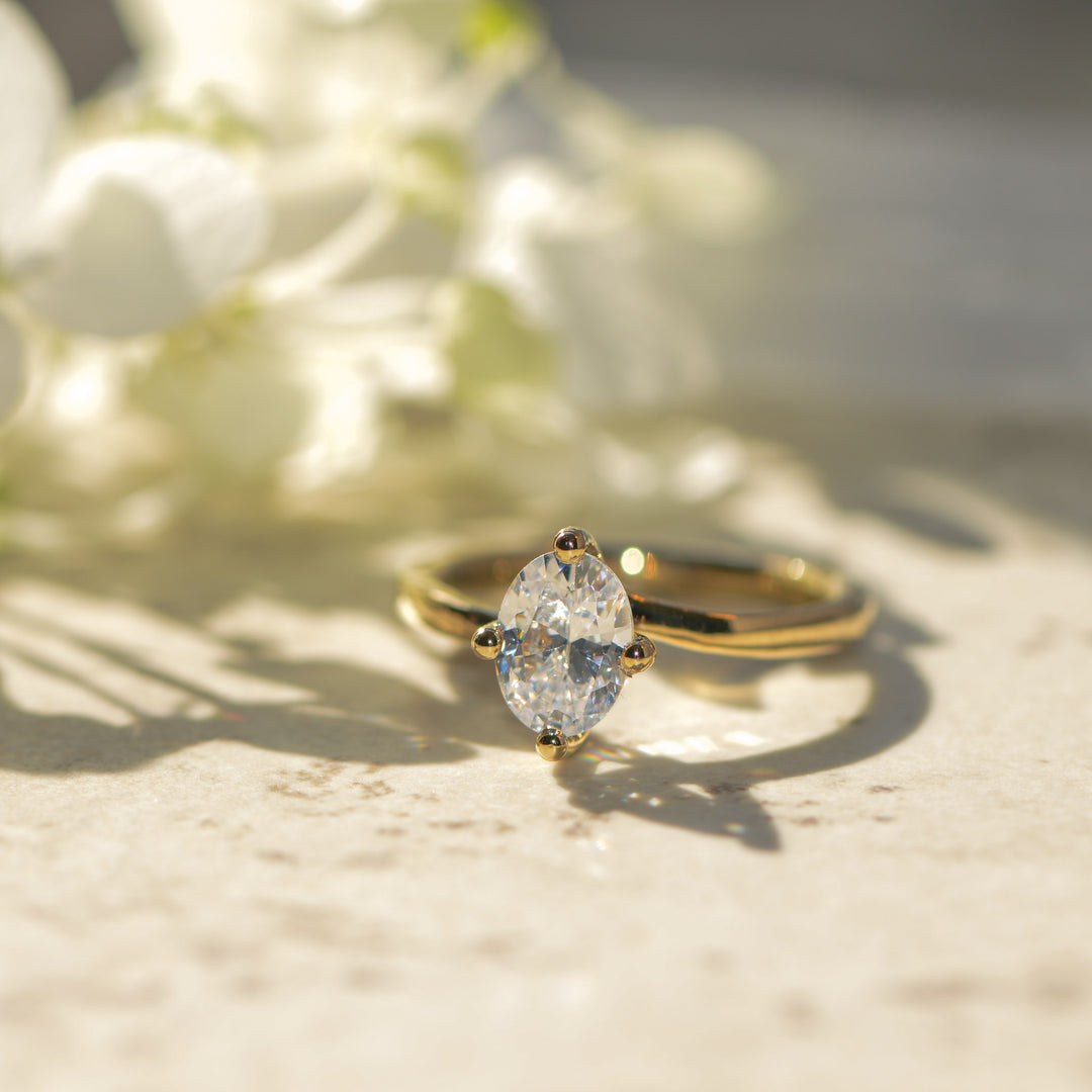 Gold engagement ring with an oval cut diamond on a light surface with blurred flowers in the background