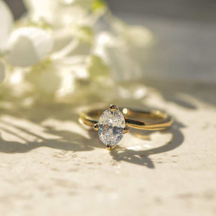 Gold engagement ring with an oval cut diamond on a light surface with blurred flowers in the background