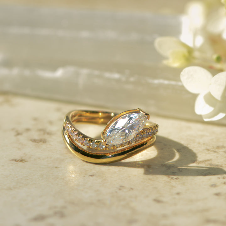 Gold pavé set engagement ring with a large marquise diamond on a marble surface with flowers in the background