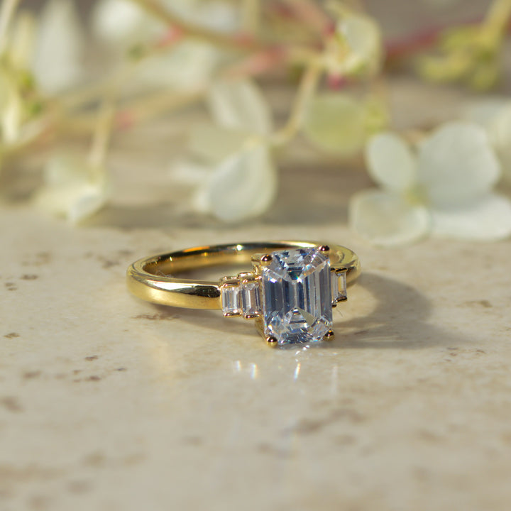Gold ring with a large emerald cut diamond centre with baguette cut side stones on a marble surface with flowers in the background