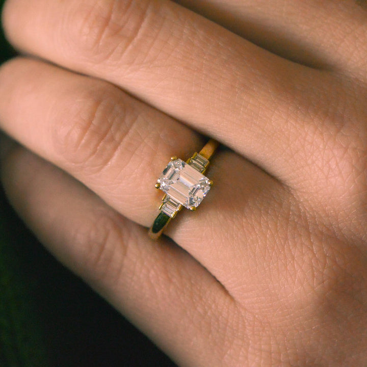 Gold art deco ring with an emerald cut diamond on a finger against a dark background