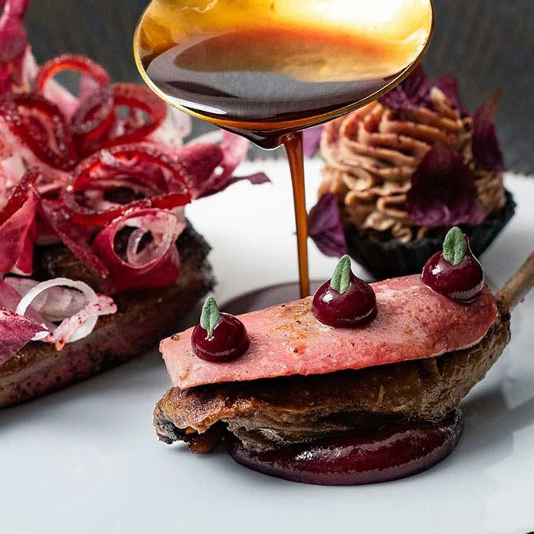 Delicate dish with pink meat, cherries, and a dark sauce being poured over it on a white plate.