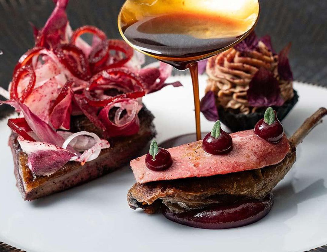 Delicate dish with red meat, onions, and a sauce being drizzled over it on a white plate.