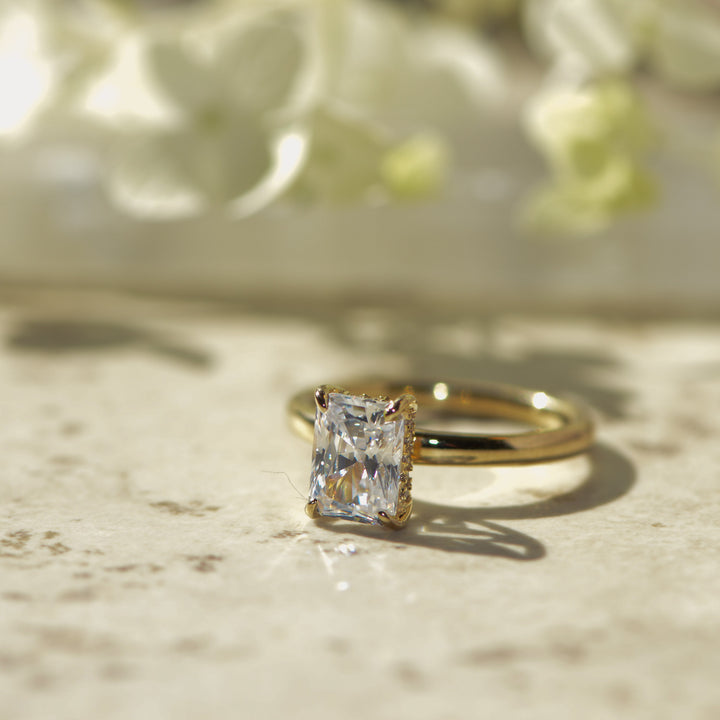 Gold ring with a large radiant diamond on a marble surface with flowers in the background