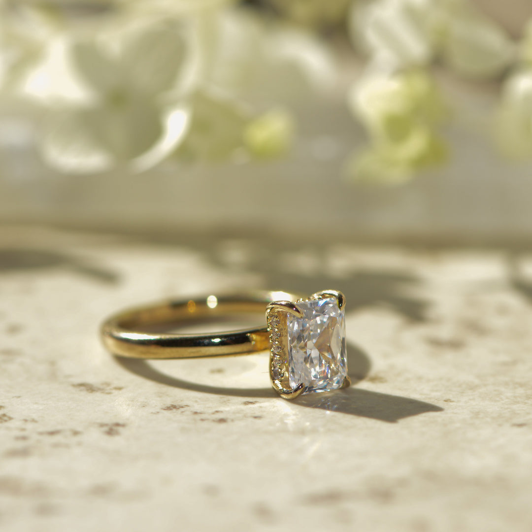 Gold ring with a radiant cut diamond on a marble surface with blurred flowers in the background