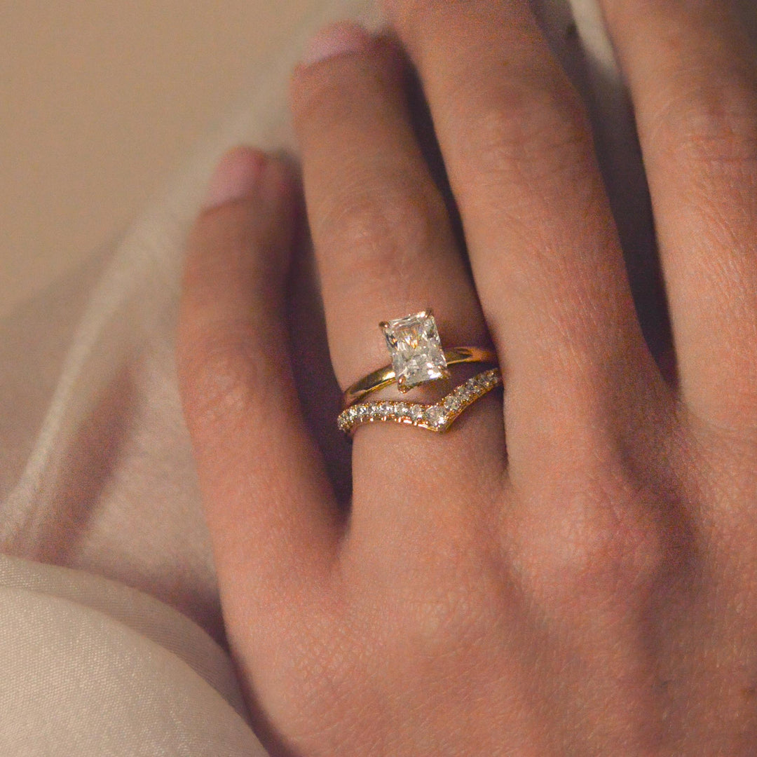 Close up of a radiant cut diamond engagement ring worn on the hand with a gold, pavé set chevron wedding band