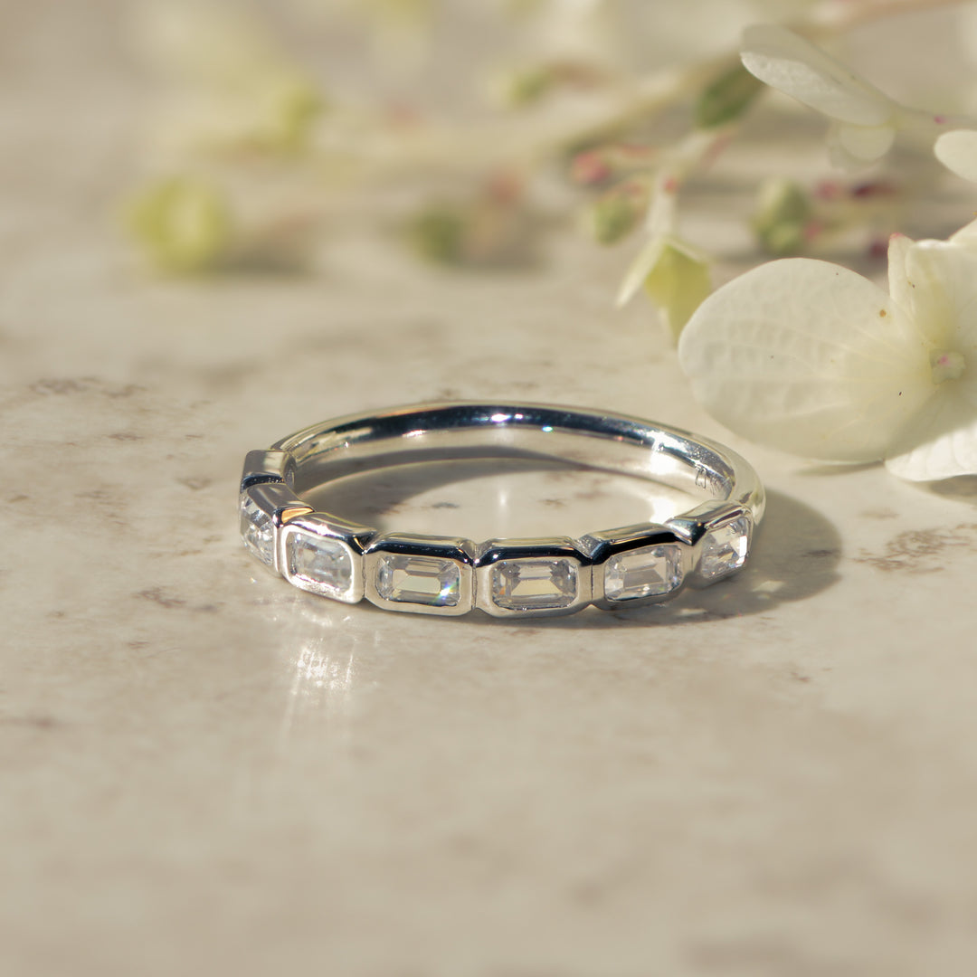 Platinum wedding ring with emerald cut diamonds on a marble surface with flowers in the background