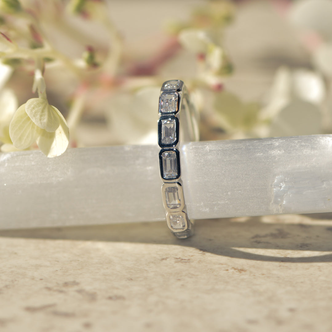Platinum ring with bezel set emerald cut diamonds on a white surface with a blurred floral background