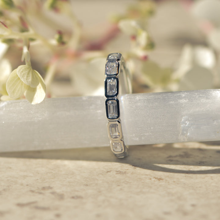 Platinum ring with bezel set emerald cut diamonds on a white surface with a blurred floral background