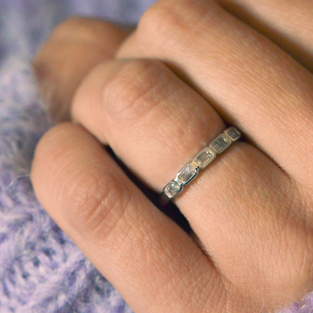 Close up of a platinum wedding band, bezel set with emerald cut diamonds worn on the hand with a purple background  