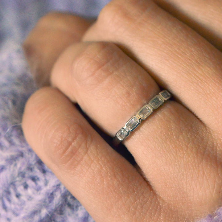 Close up of a platinum wedding band, bezel set with emerald cut diamonds worn on the hand with a purple background  
