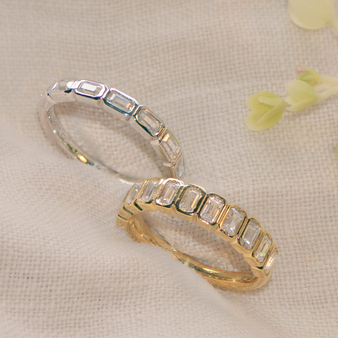 Two rings set with emerald cut diamonds, one in gold and one in platinum, on a beige linen background