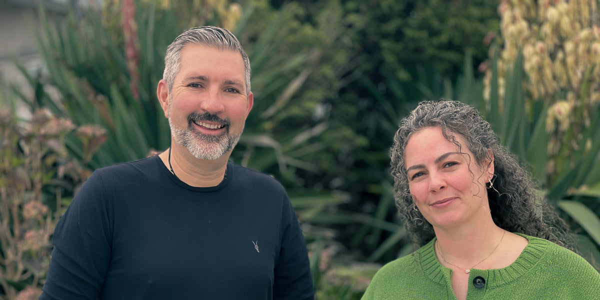 Two people standing outdoors with greenery in the background