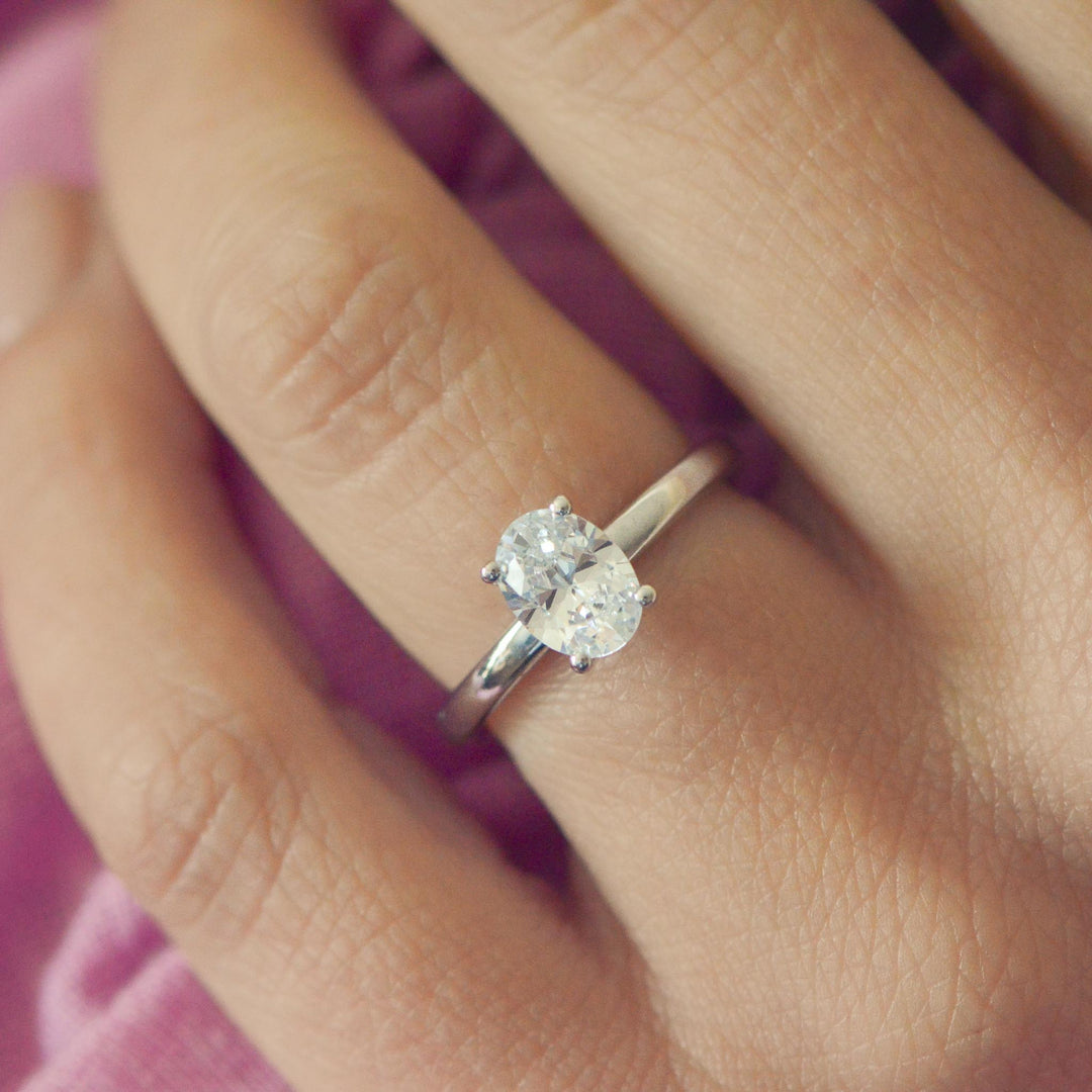 Platinum four claw solitaire ring with an oval cut diamond on a person's finger against a pink background