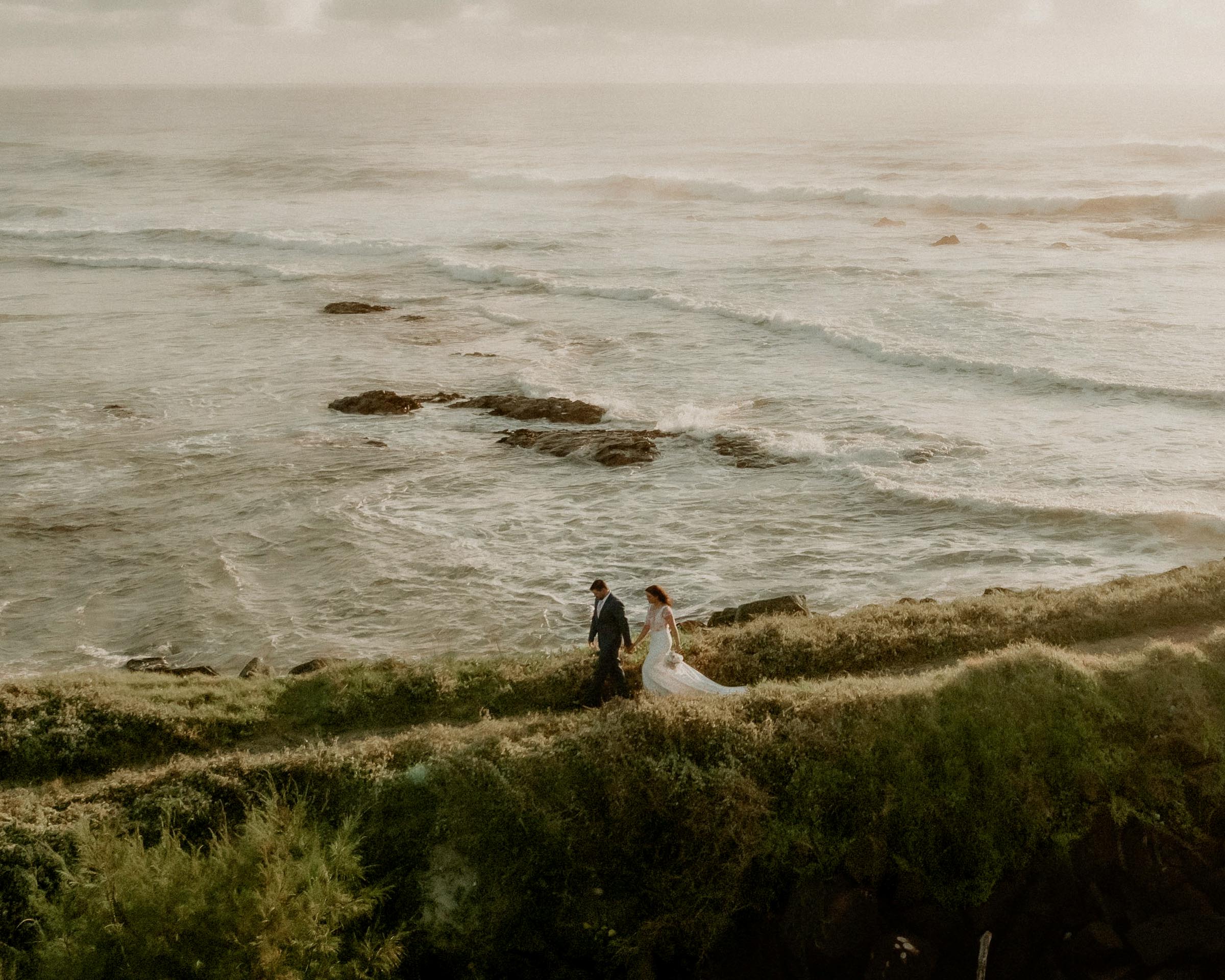 Bride and Groom walking along a grassy cliff overlooking the ocean.