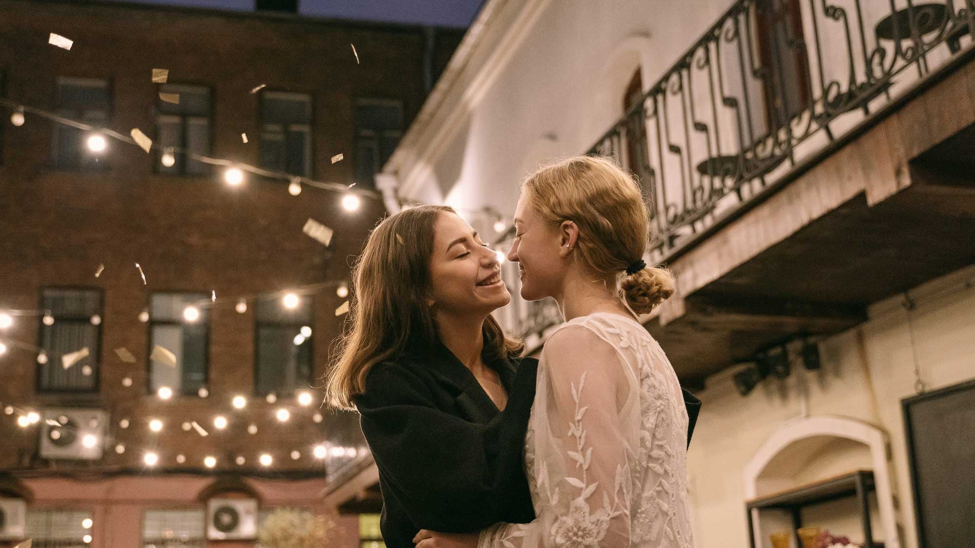 Two women embracing on a street with string lights and buildings in the background