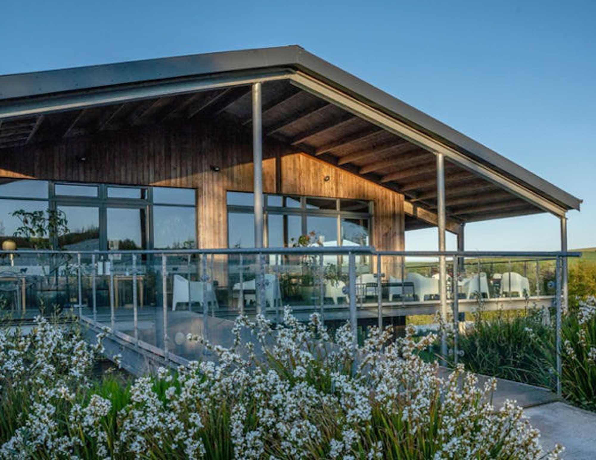 Modern wooden restaurant at Trevibban Mill with a large glass patio area, surrounded by white flowers and greenery.